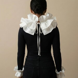 Op de afbeelding: Een zwarte top met lange mouwen en een grote witte gerimpelde kraag en bijpassende manchetten. De kraag is aan de achterkant vastgebonden met een wit lint. Het model draagt een zwarte jeans.