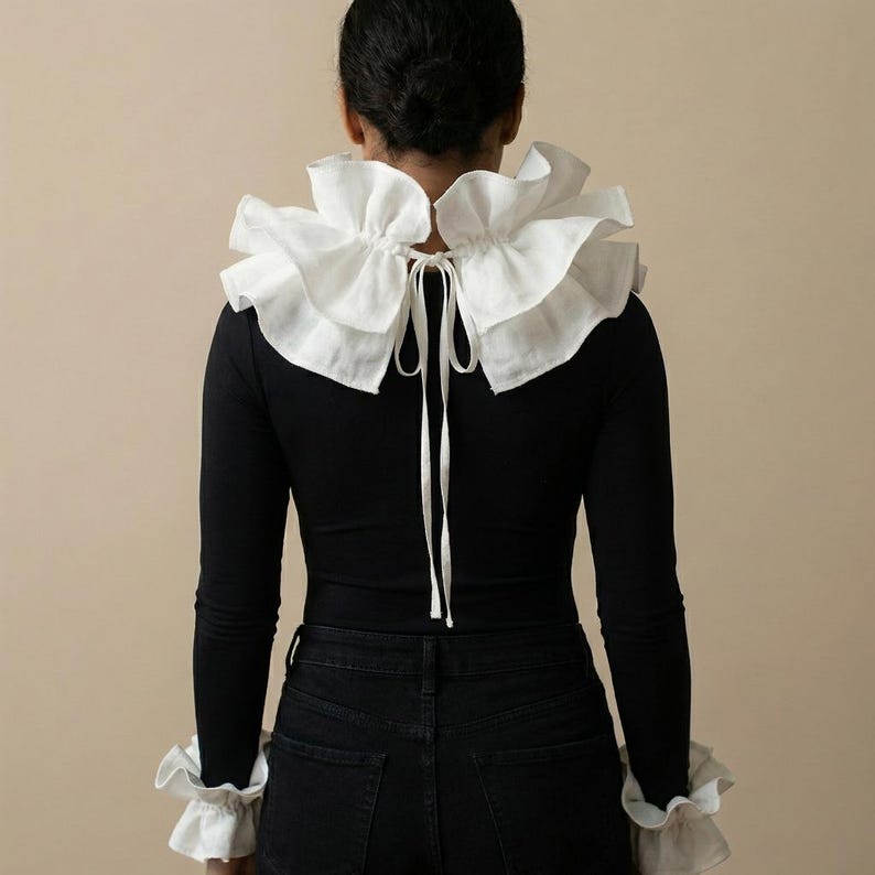 Op de afbeelding: Een zwarte top met lange mouwen en een grote witte gerimpelde kraag en bijpassende manchetten. De kraag is aan de achterkant vastgebonden met een wit lint. Het model draagt een zwarte jeans.