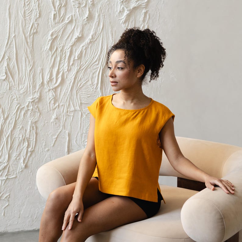 May include: Mustard yellow linen top with a round neckline and short sleeves. The top has a relaxed fit and side slits. The model is sitting on a beige chair. The background is a textured white wall.