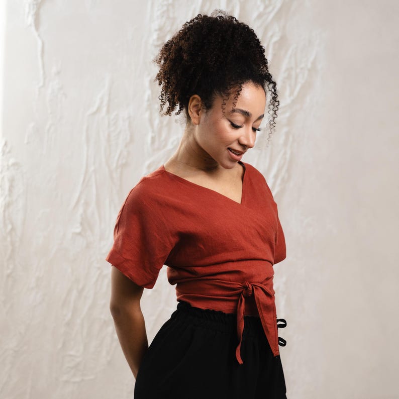 May include: A terracotta-coloured wrap top with short sleeves and a V-neck, tied at the waist. The top is worn with black trousers. The model has curly hair and is looking down.