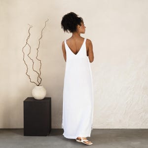 May include: A white, sleeveless maxi dress with a V-neck. The dress is floor-length and has a relaxed fit. The model is wearing white sandals. The dress is made of a lightweight fabric, perfect for warm weather.