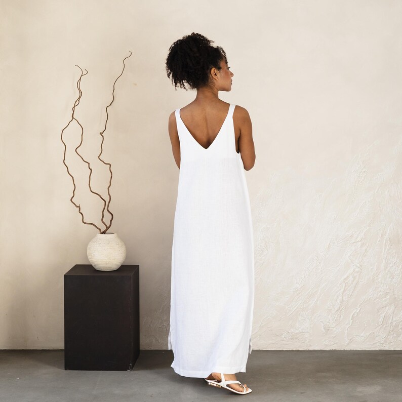 May include: A white, sleeveless maxi dress with a V-neck. The dress is floor-length and has a relaxed fit. The model is wearing white sandals. The dress is made of a lightweight fabric, perfect for warm weather.