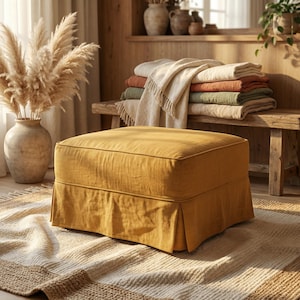 Puede incluir: Un otomán tapizado de color amarillo mostaza con una falda plisada se encuentra sobre una alfombra estampada. Un jarrón con hierba de la pampa y un banco de madera con mantas dobladas están en el fondo. La habitación tiene un ambiente cálido y acogedor.