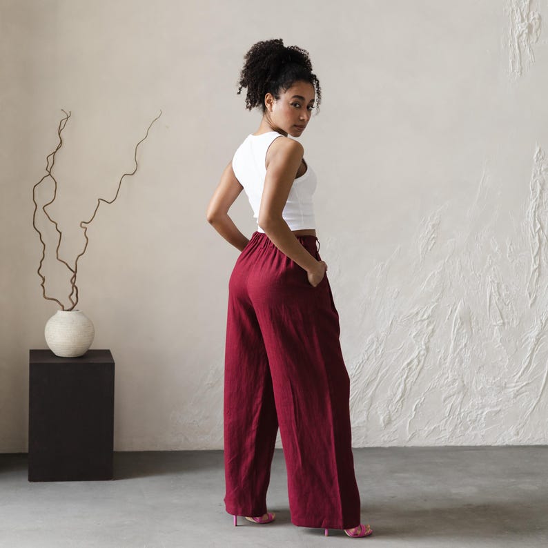 May include: A woman wearing a white tank top and wide-legged burgundy pants. The pants have a high waist and are made of a soft, flowing fabric.