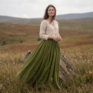 May include: A long, olive green skirt with a gathered, full silhouette paired with a beige, long-sleeved button-down shirt. The skirt reaches the ankles and features a simple waistband, while the shirt has a collar and cuffed sleeves.