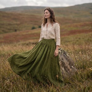 May include: A long-sleeved beige button-down shirt paired with a floor-length olive green skirt. The skirt features a gathered waistband and a full, flowing silhouette that extends to the ground, while the shirt has a simple collar and cuffs.
