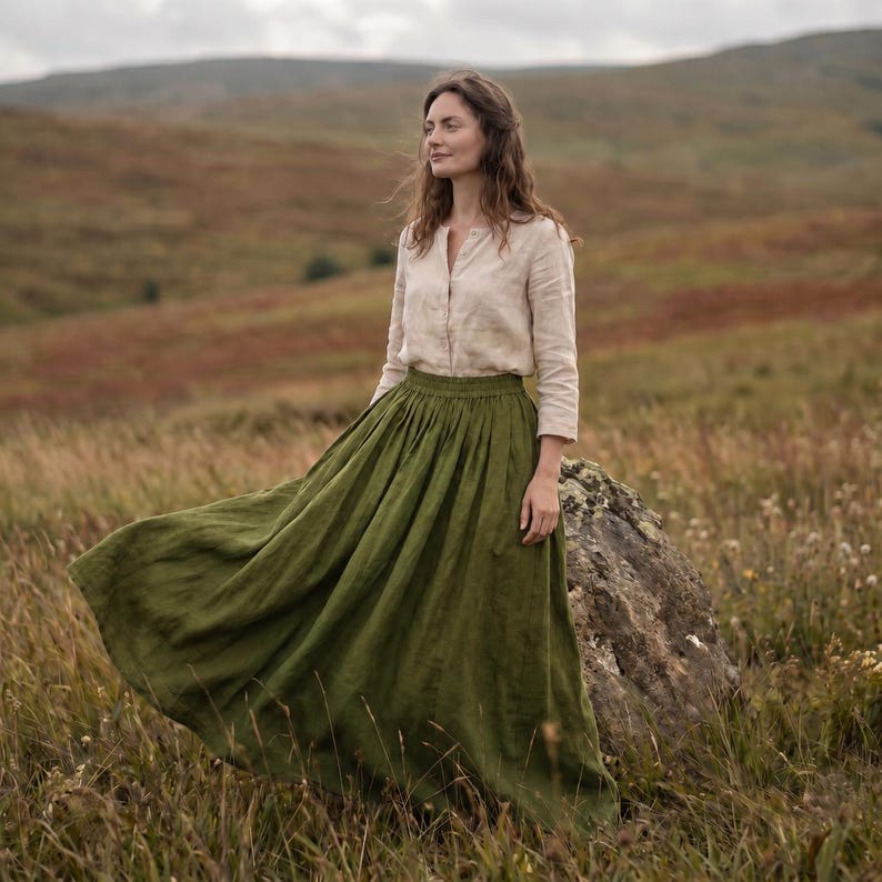 May include: A long-sleeved beige button-down shirt paired with a floor-length olive green skirt. The skirt features a gathered waistband and a full, flowing silhouette that extends to the ground, while the shirt has a simple collar and cuffs.