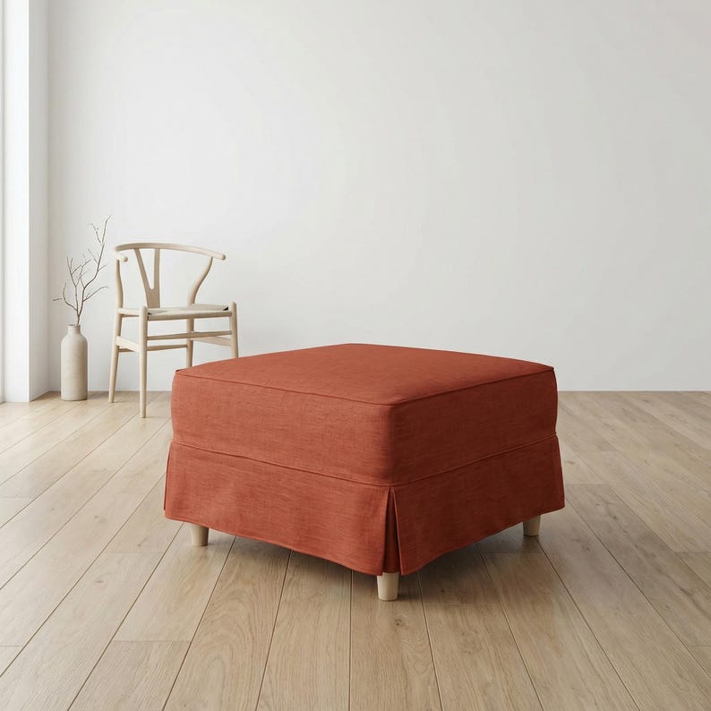May include: A square, rust-colored ottoman with a pleated skirt and light wooden legs. The ottoman is placed on a light wood floor, with a white wall and a wooden chair in the background. A vase with branches is also visible.