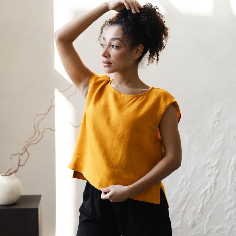 May include: Mustard yellow sleeveless top with a rounded neckline and a slightly flared hem. The top is paired with black shorts. The model is standing in front of a white textured wall.