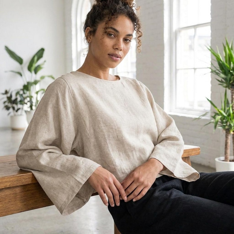 Puede incluir: Una blusa de lino beige con mangas largas y acampanadas. La blusa tiene un cuello redondo y se combina con pantalones negros. La modelo est&aacute; sentada en un banco de madera en una habitaci&oacute;n con grandes ventanas y plantas.