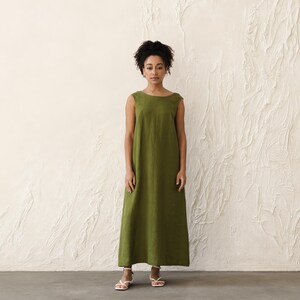 May include: A sleeveless, olive green maxi dress. The dress has a simple, round neckline and a loose, flowing fit. The model is wearing white sandals. The background is a textured, off-white wall.