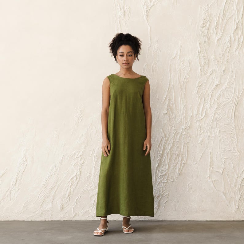 May include: A sleeveless, olive green maxi dress. The dress has a simple, round neckline and a loose, flowing fit. The model is wearing white sandals. The background is a textured, off-white wall.