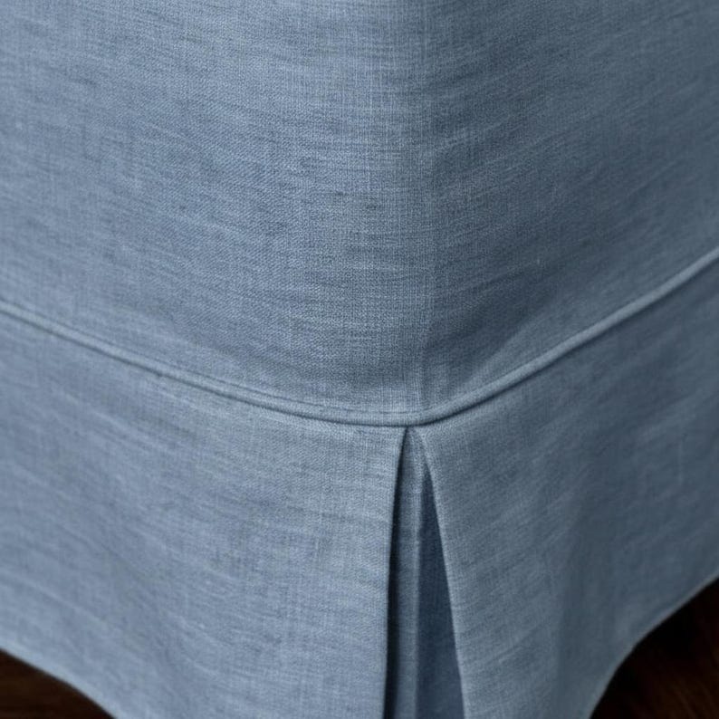 May include: Close-up of a light blue upholstered furniture corner. The fabric has a textured appearance and a subtle sheen. The corner features a tailored pleat detail, adding a touch of elegance to the design. The overall aesthetic is clean and modern.