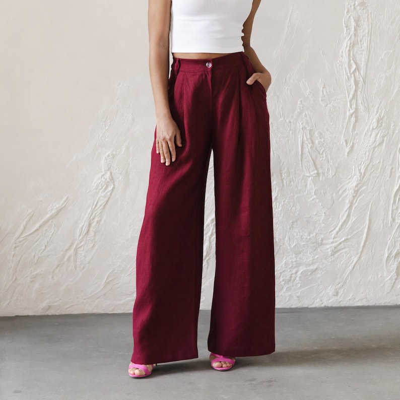 A woman is wearing high-waisted, wide-leg burgundy trousers with pleated detailing and a front button closure. She pairs the trousers with a fitted white ribbed crop top and pink heeled sandals. Her left hand rests in the pocket, and the neutral textured background complements the bold color of the pants.