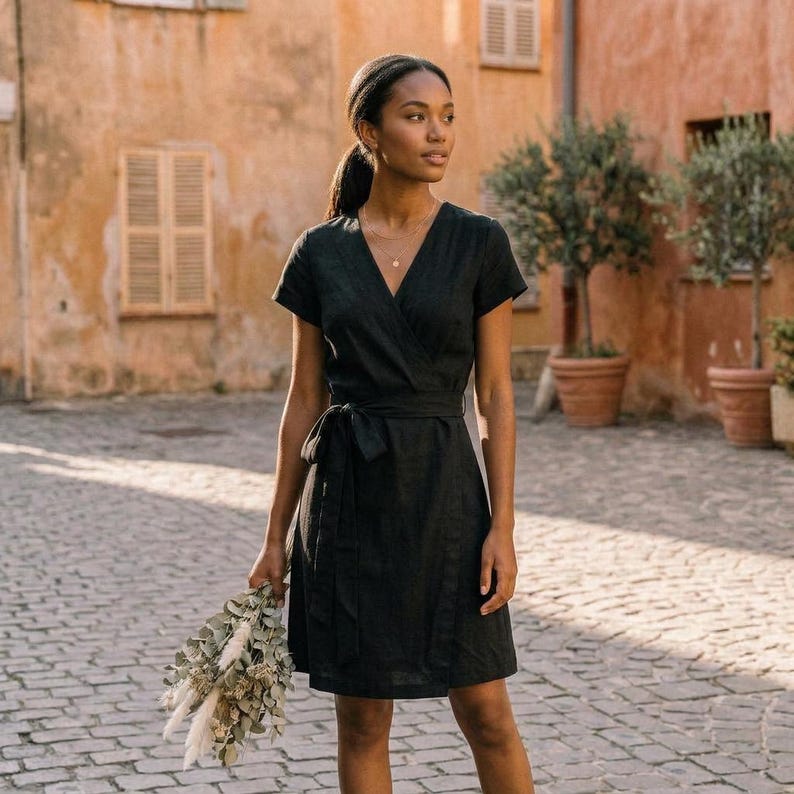 Puede incluir: Un vestido cruzado negro con mangas cortas y cintur&oacute;n a juego. El vestido est&aacute; hecho de una tela texturizada y llega a la rodilla. La persona sostiene un ramo de flores secas. El fondo presenta una calle adoquinada y edificios.