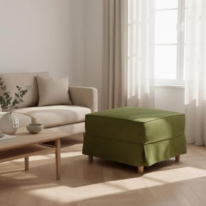 May include: An olive green upholstered ottoman with a skirted base and wooden legs. The square-shaped footrest is placed in a living room setting, near a beige sofa and a wooden coffee table.