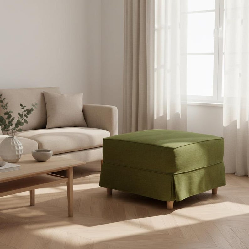 May include: An olive green upholstered ottoman with a skirted base and wooden legs. The square-shaped footrest is placed in a living room setting, near a beige sofa and a wooden coffee table.