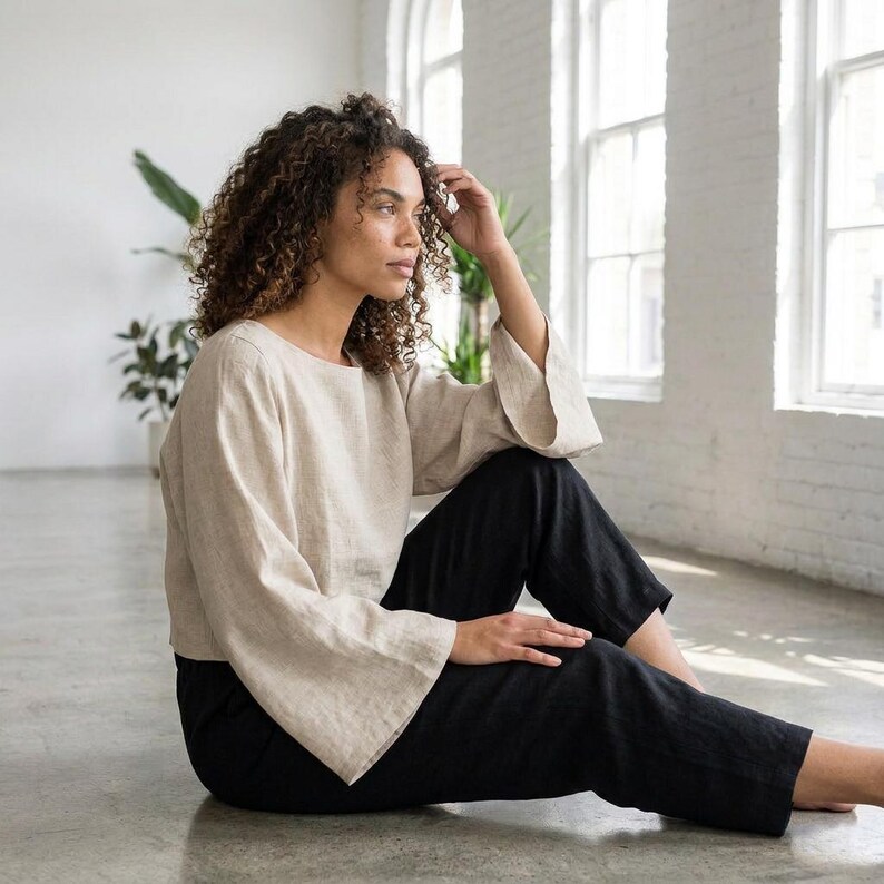 Puede incluir: Una mujer con una blusa de lino beige con mangas anchas y pantalones de lino negros. El atuendo es simple y elegante, con un ajuste relajado. La mujer est&aacute; sentada en un suelo gris claro en una habitaci&oacute;n con grandes ventanas.