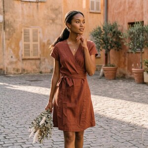 Puede incluir: Un vestido cruzado de color &oacute;xido con mangas cortas y cintura anudada. El vestido est&aacute; hecho de una tela texturizada y llega a la rodilla. La persona sostiene un ramo de flores secas. El fondo presenta una calle adoquinada y edificios.
