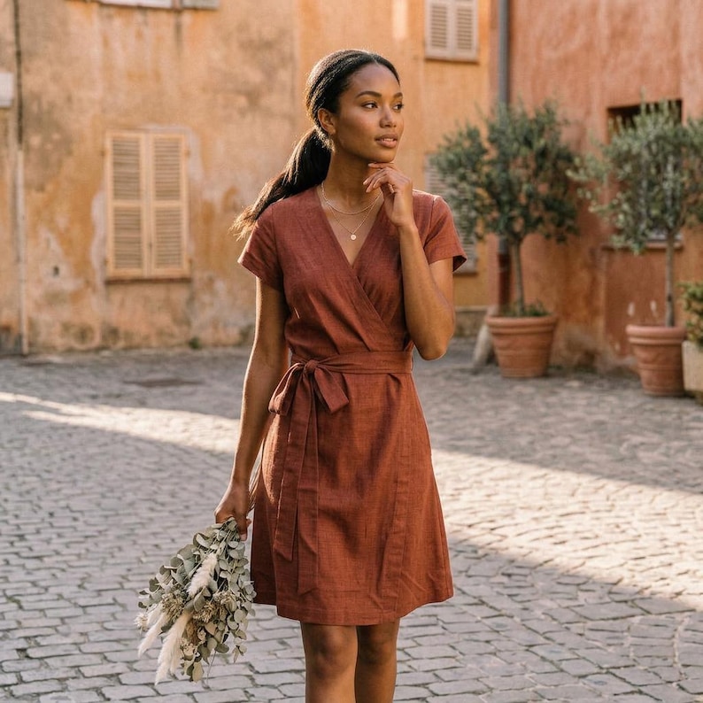 Puede incluir: Un vestido cruzado de color &oacute;xido con mangas cortas y cintura anudada. El vestido est&aacute; hecho de una tela texturizada y llega a la rodilla. La persona sostiene un ramo de flores secas. El fondo presenta una calle adoquinada y edificios.