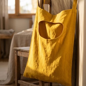 Op de afbeelding: Een mosterdgele linnen draagtas met een grote, uniek gevormde uitsparing. De tas heeft lange hengsels en lijkt gemaakt van een zachte, getextureerde stof. De tas hangt aan een houten stoel.