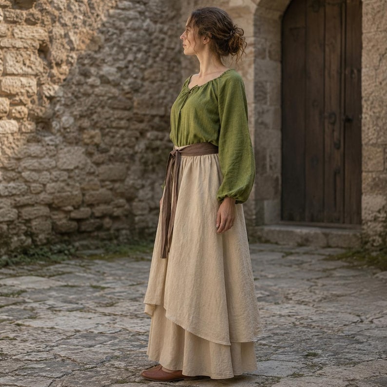 May include: A long-sleeved olive green blouse with a gathered neckline paired with a floor-length beige skirt featuring a layered hem and a brown fabric belt tied at the waist. The outfit consists of a solid-colored top and a neutral-toned skirt.