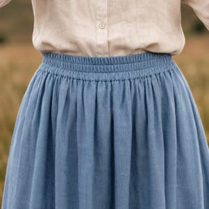 May include: A blue skirt with an elasticated waistband featuring gathered fabric details, paired with a beige button-down shirt. The skirt has a textured appearance and falls in soft vertical folds from the waistline down to the hemline.