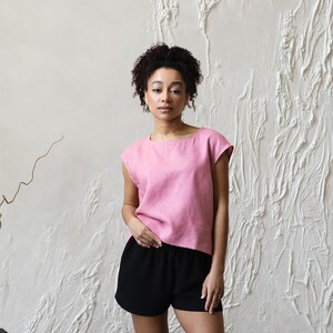 May include: A pink linen top with a boat neck and cap sleeves paired with black shorts. The outfit is simple and casual, suitable for warm weather. The background is a textured white wall.
