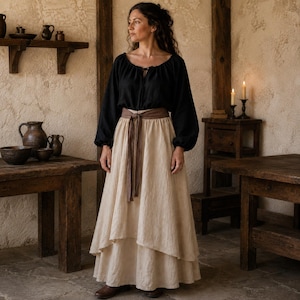 May include: A black long-sleeved blouse with a gathered neckline and tie closure paired with a floor-length beige layered skirt featuring a brown fabric belt tied at the waist. The skirt has a high-low hemline and a textured appearance.