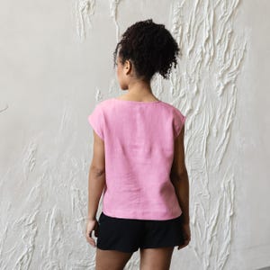 May include: A light pink linen top with a scoop neck and short sleeves. The top is paired with black shorts. The model is facing away from the camera, with their hair pulled back. The background is a textured white wall.