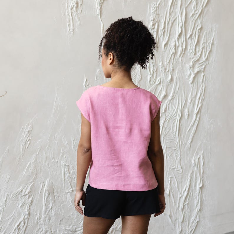May include: A light pink linen top with a scoop neck and short sleeves. The top is paired with black shorts. The model is facing away from the camera, with their hair pulled back. The background is a textured white wall.