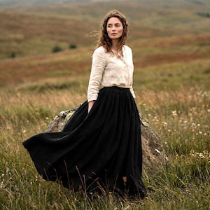 May include: A long-sleeved, light beige button-down shirt paired with a floor-length, solid black skirt. The shirt features a collared neckline and cuffed sleeves, while the skirt has a full, gathered silhouette that flows towards the ground.