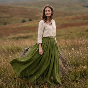 May include: A long, olive green skirt with a gathered, high-waisted design and a full, flowing silhouette, paired with a long-sleeved, light beige button-down shirt featuring a collar and a simple, straight-cut hemline.