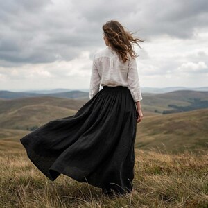 May include: A long, flowing black skirt paired with a white, long-sleeved button-down shirt. The skirt features a high waist and a full, pleated silhouette that extends to the ankles, while the shirt has a collar and cuffed sleeves.