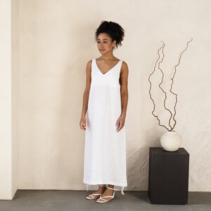 May include: A sleeveless, white V-neck maxi dress. The dress is floor-length and has a simple, elegant design. The model is wearing white sandals. The background is a neutral color, with a black square pedestal and a vase with branches.