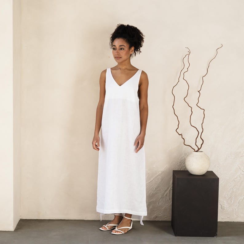 May include: A sleeveless, white V-neck maxi dress. The dress is floor-length and has a simple, elegant design. The model is wearing white sandals. The background is a neutral color, with a black square pedestal and a vase with branches.