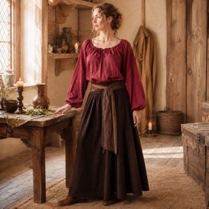 May include: A long-sleeved dark red blouse with a gathered neckline and tie detail, paired with a floor-length dark brown skirt featuring a wide, wrap-around fabric belt tied at the waist. The outfit is made of a textured, woven fabric.
