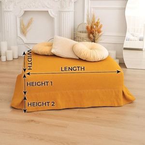 May include: Mustard yellow ottoman with a pleated skirt, topped with three decorative pillows in beige and mustard yellow. The ottoman is rectangular and appears to be made of a soft fabric. The image also shows a white fireplace and a mirror.