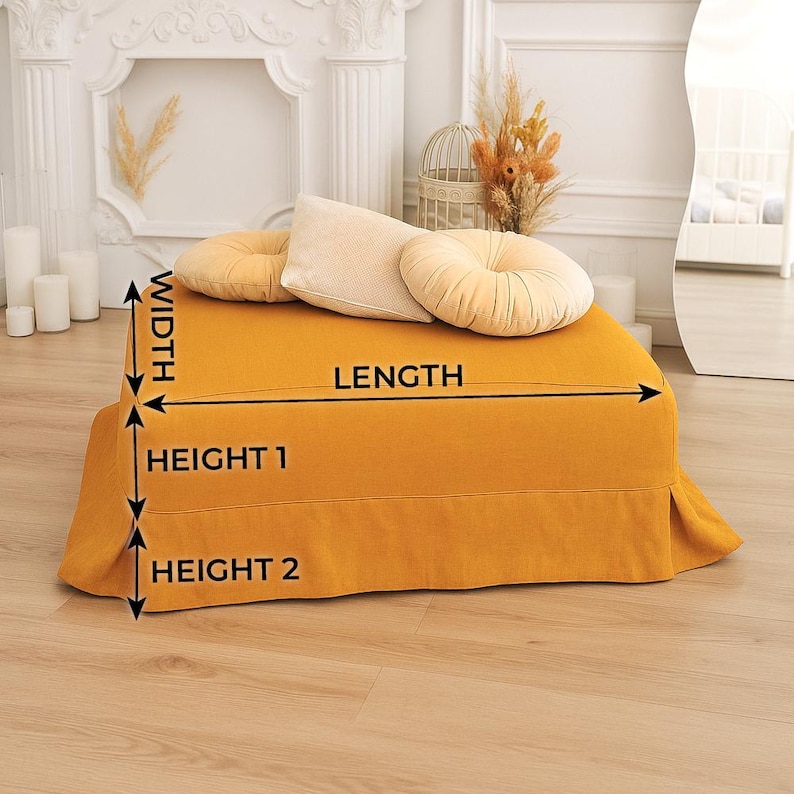May include: Mustard yellow ottoman with a pleated skirt, topped with three decorative pillows in beige and mustard yellow. The ottoman is rectangular and appears to be made of a soft fabric. The image also shows a white fireplace and a mirror.