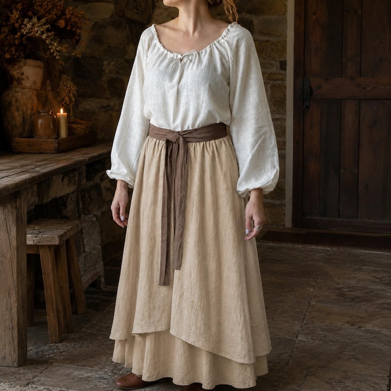 May include: A long-sleeved white blouse with a gathered neckline and drawstring tie paired with a floor-length beige layered skirt. A wide brown fabric belt is tied around the waist, securing the skirt with a visible knot and long hanging ends.