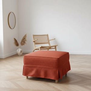 May include: An orange-red upholstered ottoman with a skirted base and wooden legs. The square-shaped footstool is in a room with a wooden chair, a vase with dried plants, and a round mirror.