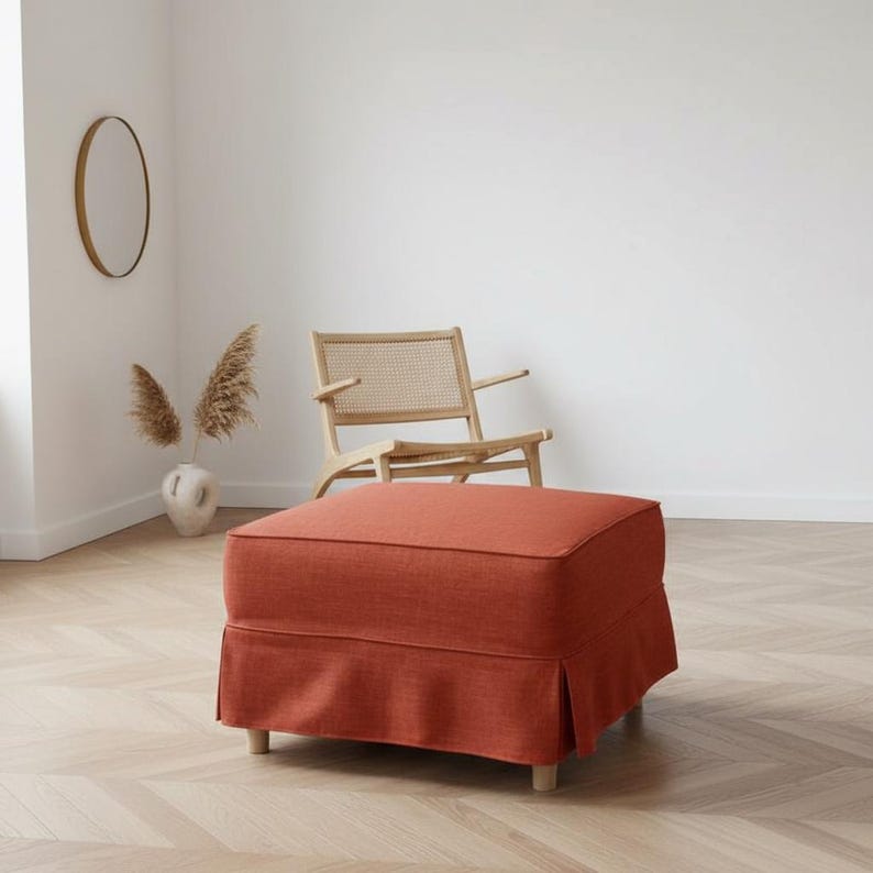 May include: An orange-red upholstered ottoman with a skirted base and wooden legs. The square-shaped footstool is in a room with a wooden chair, a vase with dried plants, and a round mirror.