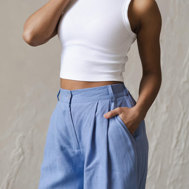 May include: A white sleeveless crop top paired with light blue high-waisted pants. The pants have pleats and a button closure.