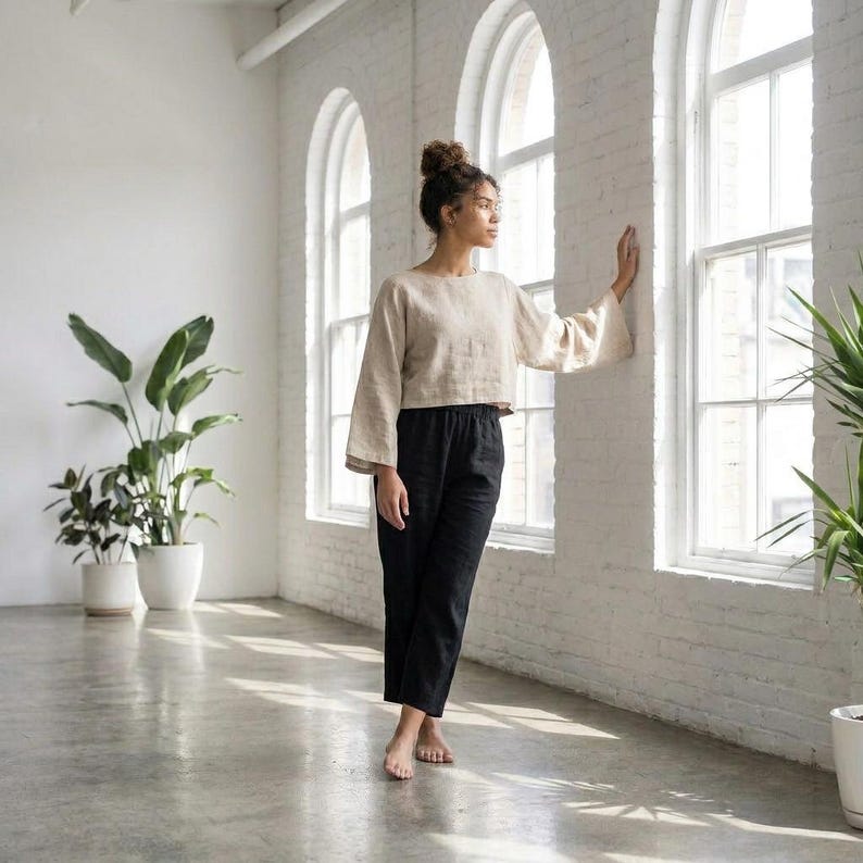 Puede incluir: Una mujer con una blusa de lino beige con mangas anchas y pantalones de lino negros. El atuendo es sencillo y elegante. Est&aacute; de pie en una habitaci&oacute;n luminosa con grandes ventanas y plantas.