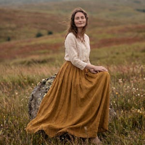 May include: A long-sleeved beige button-down shirt paired with a floor-length mustard yellow skirt. The skirt features a gathered waistline and a full, flowing silhouette that drapes over a large rock in an open, grassy field landscape.