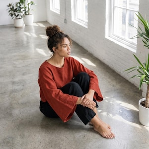 Puede incluir: Una blusa de manga larga color &oacute;xido con mangas anchas y pantalones negros. La persona est&aacute; sentada en un suelo de hormig&oacute;n cerca de una ventana y plantas en macetas. La blusa tiene un cuello redondo.