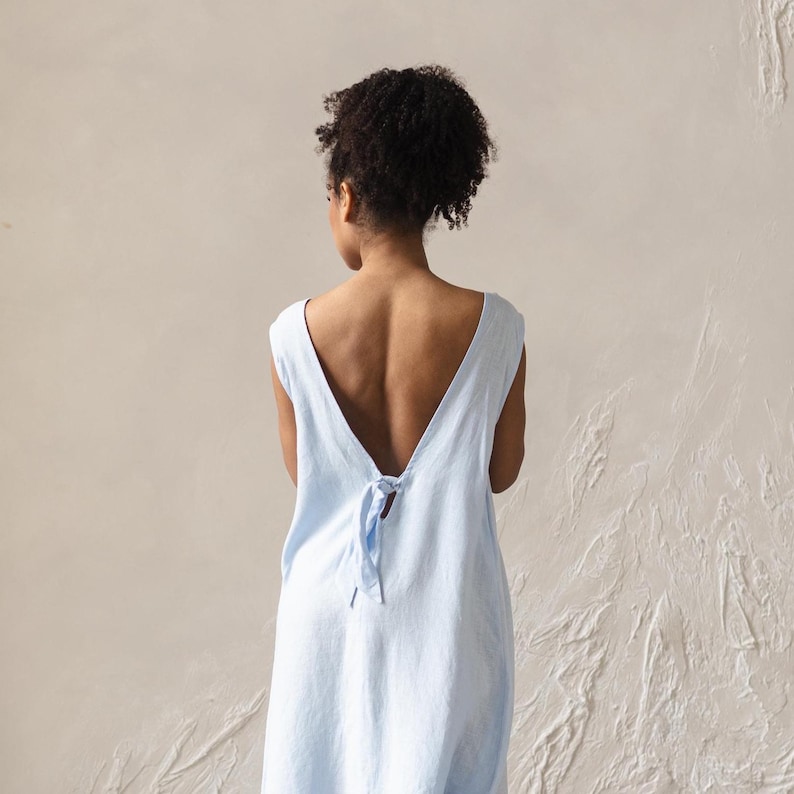 May include: A light blue sleeveless dress with a deep V-back and a tie closure. The dress is made of a soft, flowing fabric and has a simple, elegant design. The dress is a solid color and appears to be a linen or cotton blend.