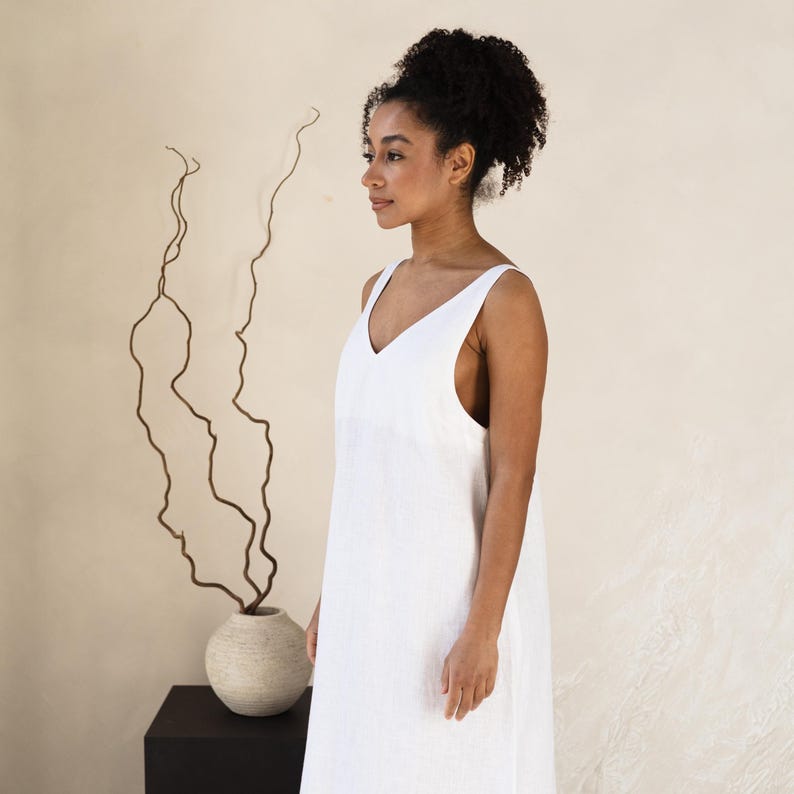 May include: A white, sleeveless V-neck dress. The dress is made of a lightweight fabric and falls to the mid-calf. A woman is wearing the dress. A decorative vase with branches is in the background.