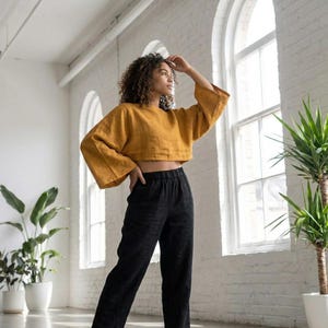 Puede incluir: Top corto amarillo mostaza con mangas anchas y pantalones de lino negros. El conjunto est&aacute; hecho de una tela suave y transpirable. Los pantalones tienen cintura el&aacute;stica. La modelo est&aacute; de pie frente a una pared de ladrillos blancos y grandes ventanas.