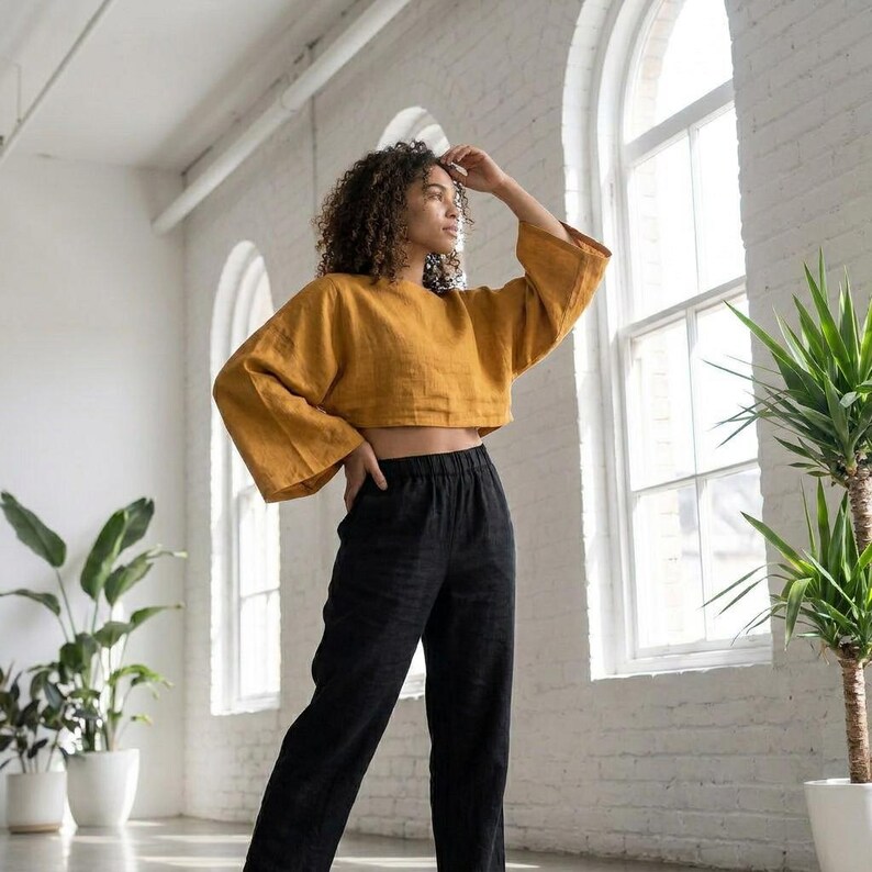 Puede incluir: Top corto amarillo mostaza con mangas anchas y pantalones de lino negros. El conjunto est&aacute; hecho de una tela suave y transpirable. Los pantalones tienen cintura el&aacute;stica. La modelo est&aacute; de pie frente a una pared de ladrillos blancos y grandes ventanas.